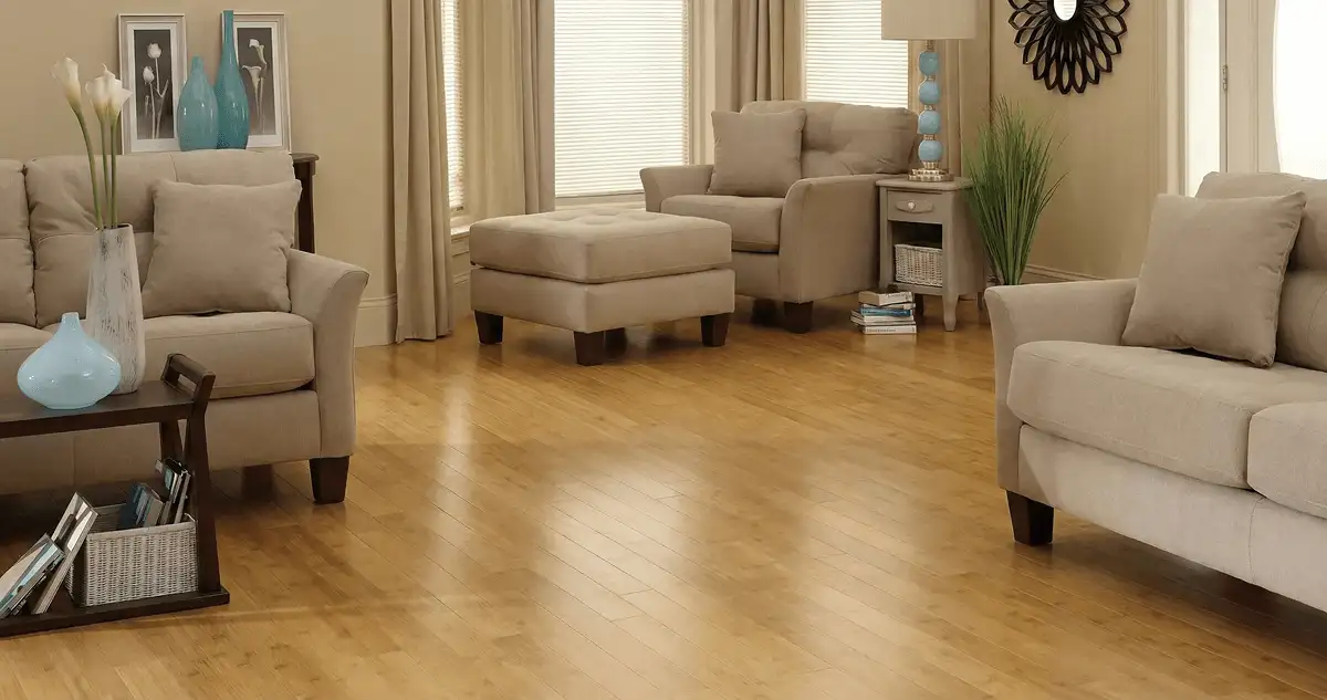 A living room with professionally installed bamboo flooring, beige sofas, a matching ottoman, a side table with a blue lamp, vases, framed pictures, and large windows with blinds and curtains in Denver, CO